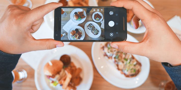 Las manos de una persona sosteniendo un teléfono celular para tomar una foto a varios platos con alimentos en una empresa de food tech.