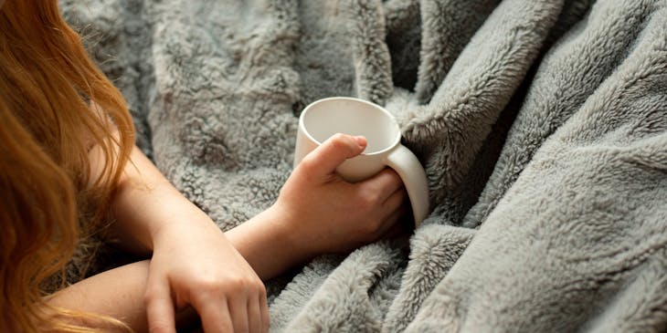 Mujer con una taza de café en la mano, tapada con una cobija de color gris en un negocio de cobijas.