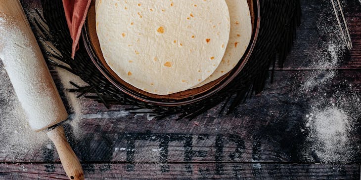 Tortillero lleno de tortillas de harina sobre una mesa de madera con harina esparcida, rodeado de un palote y una cuchara de medir en un negocio de tortillas de harina.