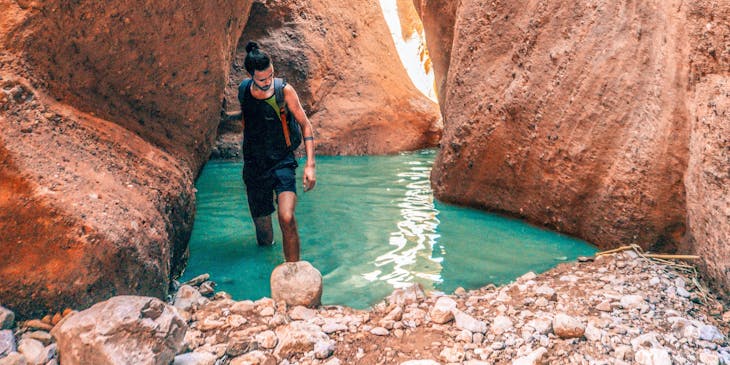 Man hiking through blue water between tall canyon walls.