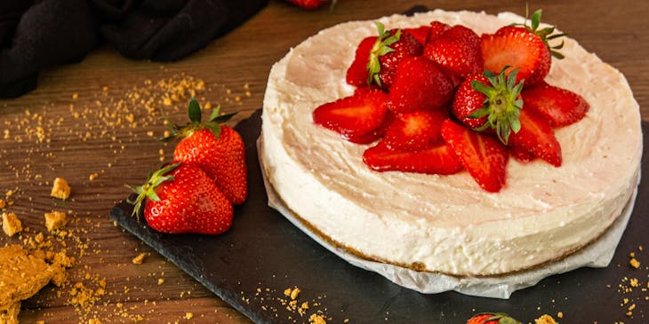 Una cheesecake decorata con fragole servita su un piatto nero sopra un tavolo di legno.