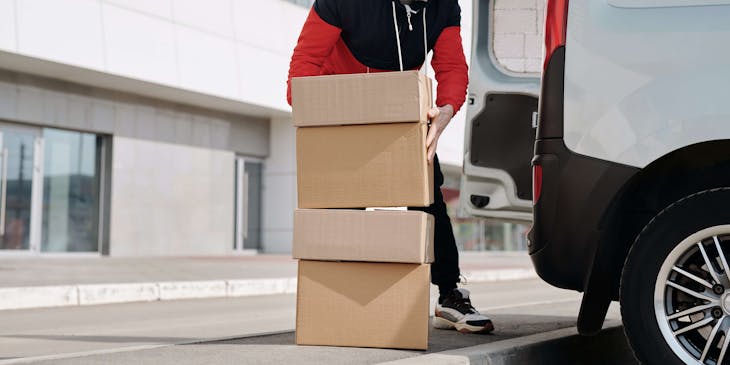 Un mensajero con un par de cajas colocándolas en la parte trasera de una camioneta blanca en un negocio de servicios de mensajería y transporte.