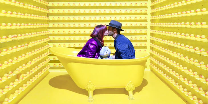 Duas pessoas se beijando em uma banheira amarela cercada por patos de borracha amarelos.