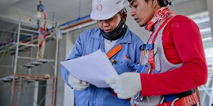 Dos ingenieros con cascos y equipo de seguridad mirando planos en una empresa constructora.