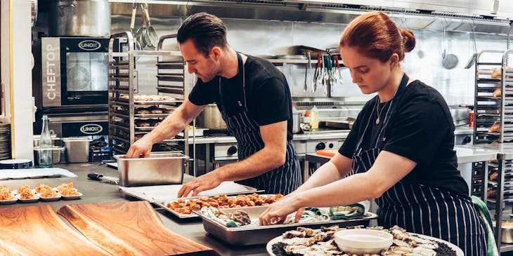 Dois chefs vestidos com aventais e preparando pratos de comida em uma cozinha compartilhada.