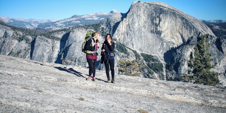 Dos mujeres con mochilas en la espalda caminando en una montaña y un ave volando al fondo en una empresa de turismo de aventura.