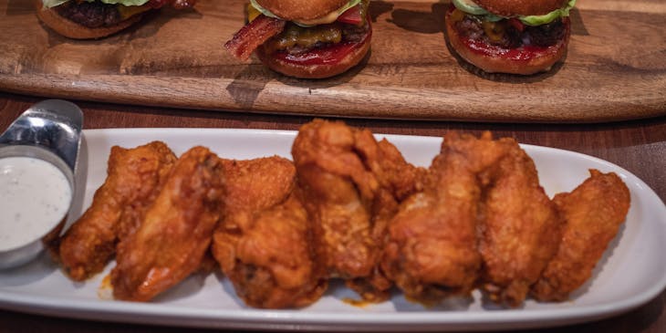 Un plato de cerámica blanca lleno de alitas de pollo picantes frente a una bandeja de madera llena de hamburguesas en un restaurante de alitas y hamburguesas.