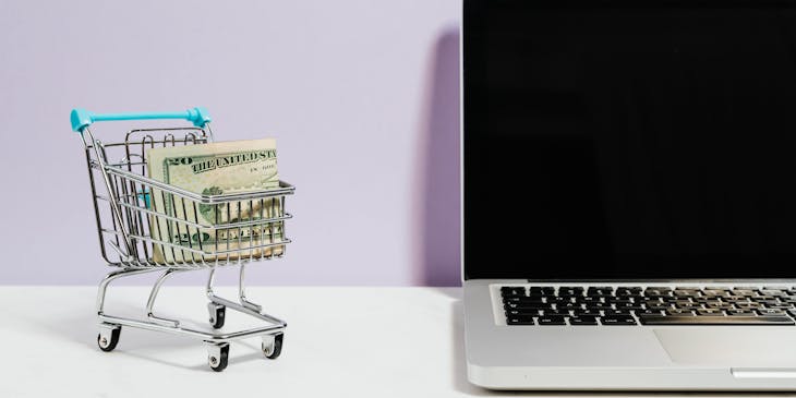 La imagen de un carrito de compras miniatura con billetes dentro frente a una laptop en una empresa de e-commerce.