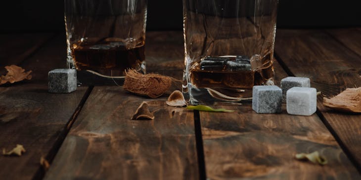 Dos vasos servidos con ron de una empresa de ron venezolano sobre madera con hojas secas esparcidas y cubos de azúcar.