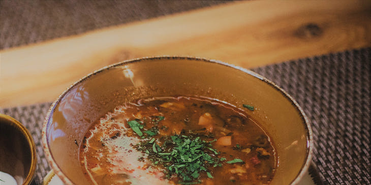 Un plato con menudo a un lado de una cuchara y un vaso pequeño con agua de horchata sobre un mantel en una mesa de una menudería.