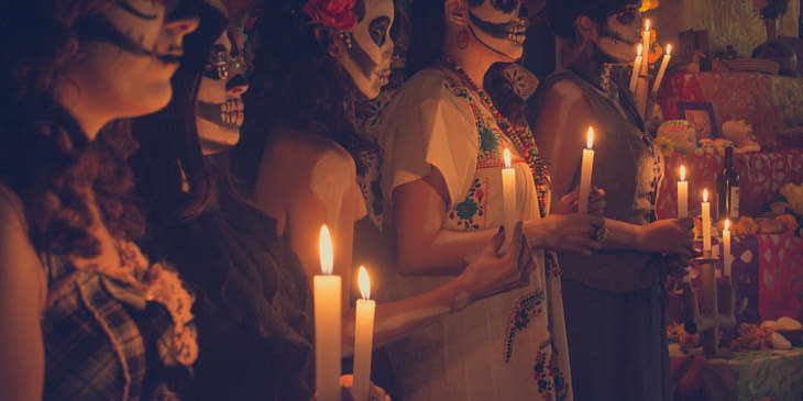 Un grupo de mujeres caracterizadas como catrinas sosteniendo velas frente a un altar de un negocio de Día de Muertos