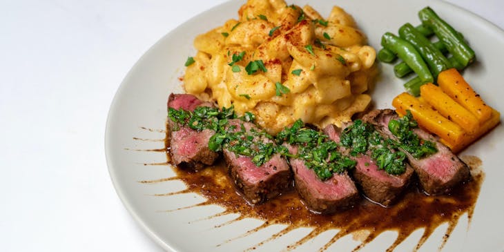 Un corte de carne a termino medio y mac and cheese con chimichurri encima servido en un plato.