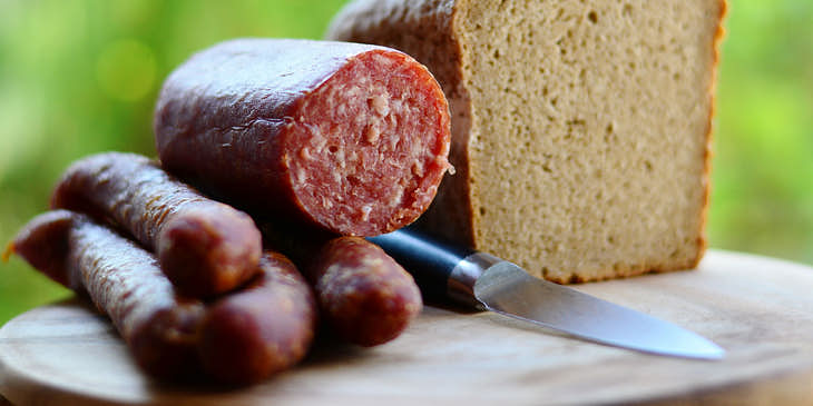 Unos trozos de chorizo a un lado de una pieza de pan blanco y un cuchillo sobre una tabla de madera para picar fuera de un negocio de choripán.