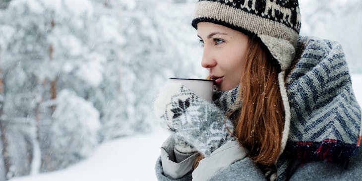 Una mujer en un bosque nevado tomando café usando un gorro de un negocio de chullos.