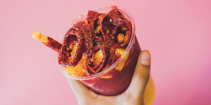 Mango picado con chamoy en un vaso transparente sujetado por una mano con un fondo rosa.