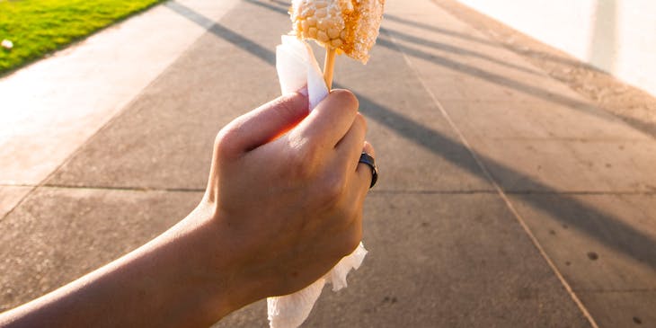 Persona sujetando un elote de un negocio de elote loco en un camino rodeado por palmeras y un edificio al lado.