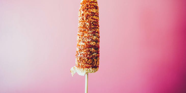 Un elote con mayonesa y chile en polvo con queso rallado espolvoreado sobre un fondo color rosa.