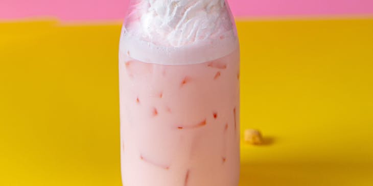 Una malteada de fresa con crema batida encima y trozos de galleta frente a un fondo rosa y amarillo en un negocio de malteadas.