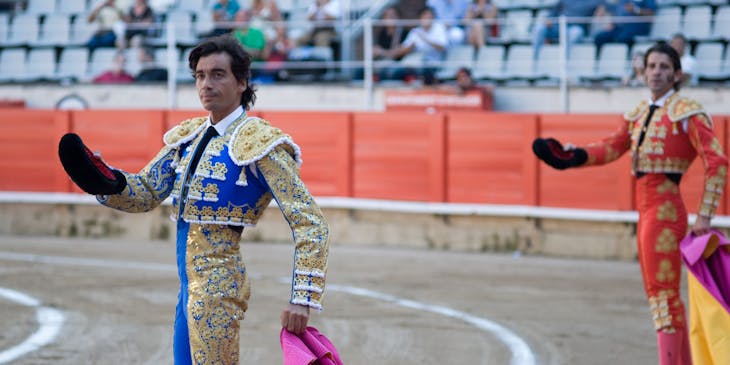 Dos toreros en el ruedo vistiendo el traje de luces color azul y color rojo respectivamente con público al fondo.