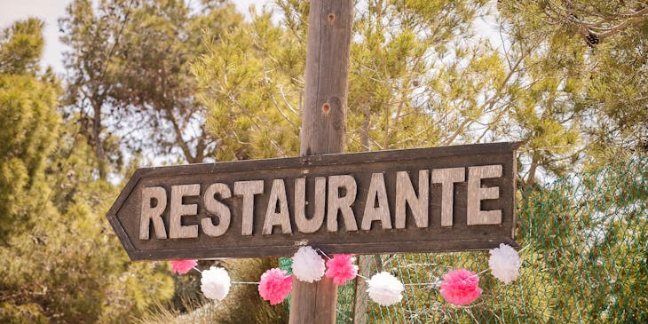 Señalamiento de un restaurante campestre colgado de un poste de madera entre árboles y flores de papel.