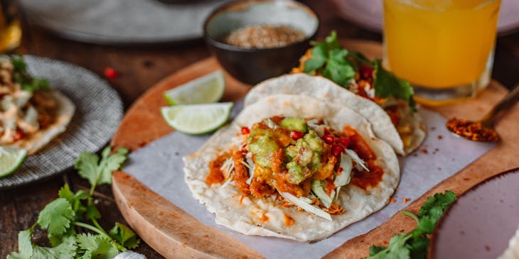 Unos tacos de cochinita sobre una base de madera con una bebida refrescante al lado limones y otros platos en el fondo en un restaurante de cochinita.