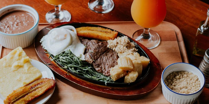 Tres vasos de jugos a un lado de un platillo de carne, verduras y otro con un plátano y pan en un restaurante de minutas.