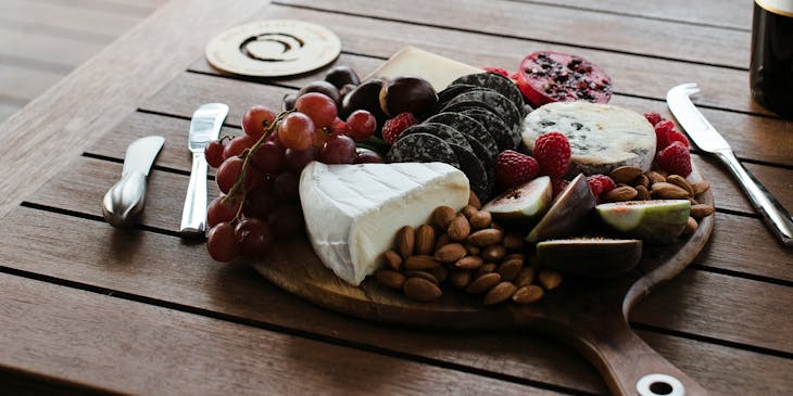 Una bandeja de madera con fruta, quesos, fiambres, y almendras a un lado de una botella de vino y cubiertos sobre una mesa en un restaurante de picoteo.