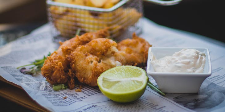 Pescado frito sobre un periódico con medio limón, aderezo y una canastilla de papas al aceite en un restaurante inglés.