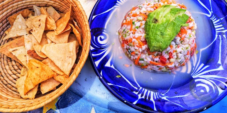 Un plato azul servido con ceviche y una canasta con totopos sobre una mesa decorada con un sol en un restaurante peruano.