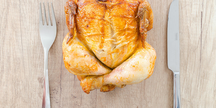 Un pollo rostizado en una mesa de madera con dos cubiertos a un lado en una rosticería.