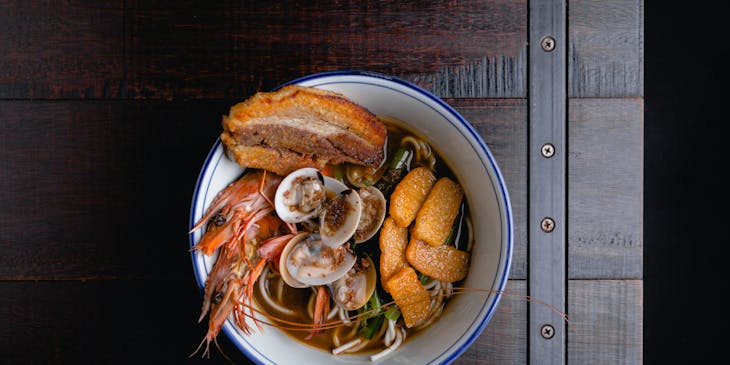 White bowl of noodles in broth with a variety of seafood on a dark wooden table at a Singaporean restaurant.