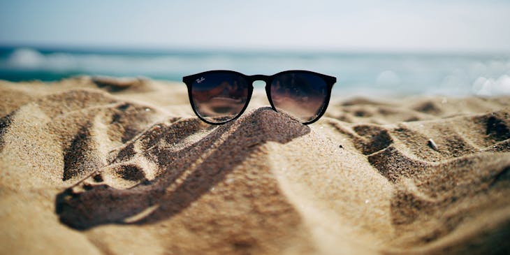 Eine Sonnenbrille liegt auf einem Sandstrand mit dem Meer im Hintergrund.