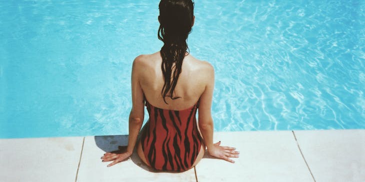 Eine Frau präsentiert sitzend am Beckenrad eines Swimmingpools ihren neuen Badeanzug, den Sie in einem Bademodengeschäft gekauft hat, und hält die Füße ins Wasser.