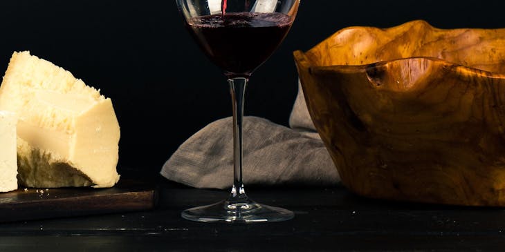 Una persona sirviendo una copa de vino tinto sobre una mesa con un plato de quesos en un bar de vino.